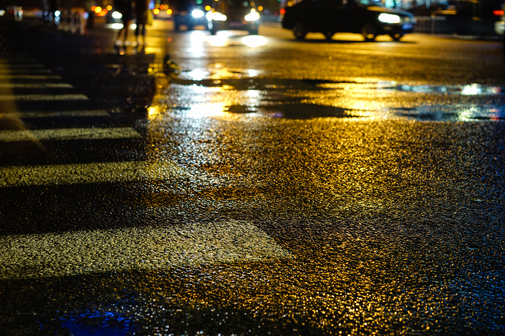 Rainy night in the big city, the light from the headlamps of vehicles approaching on the road. Close up view from the level of asphalt, focus on the asphalt