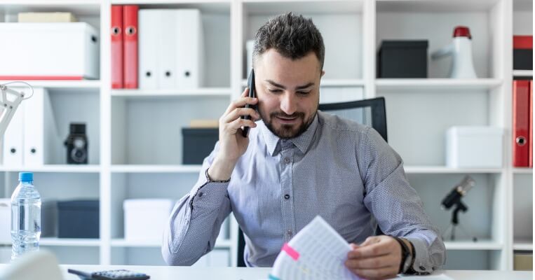 Man looking at papers on desk while on the phone, he is smiling.