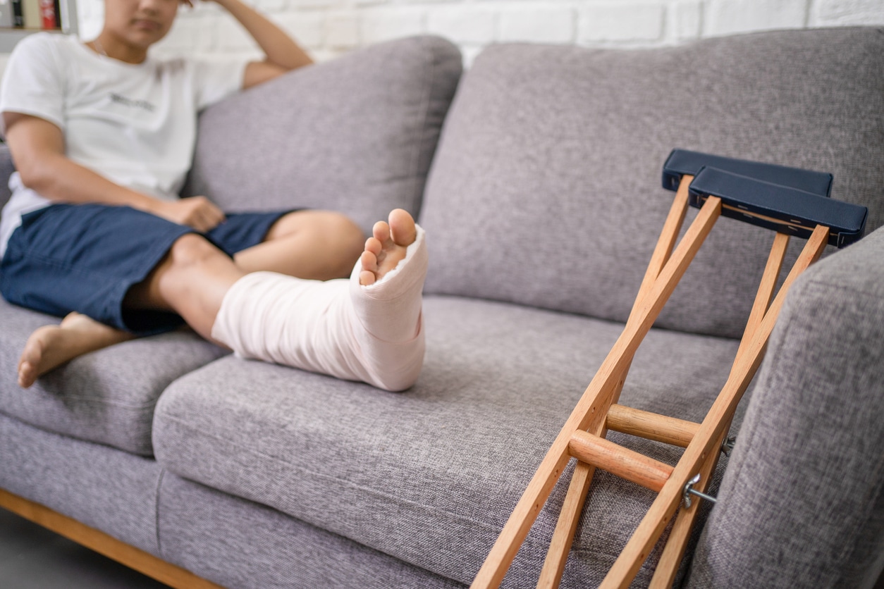 Young Asian man with a broken leg sitting on the sofa instead of working. Close up of the crutch.